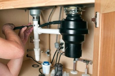 Plumber repairing a garbage disposal beneath a kitchen sink in Carlisle